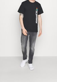 Camiseta de algodón negra con diseño triangular colorido y texto blanco en el costado, combinada con jeans gris ajustados y zapatillas blancas.