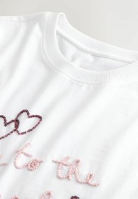 Tricou alb cu guler rotund, cu text roz scris în stil cursiv din mărgele și două inimi roșii din mărgele suprapuse în zona pieptului.