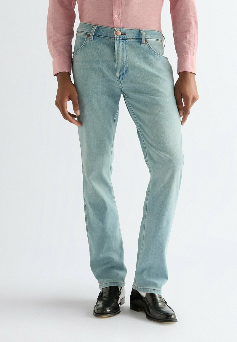 Homme portant un jean slim bleu clair, des mocassins en cuir noirs et une chemise rose boutonnée, debout devant un fond uni.