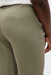 Close-up van een persoon die olijfgroene broek draagt met een achterzak met klep en een wit overhemd in de broek gestopt, met zicht op de zijkant en heupgebied.