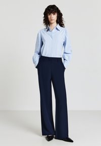 Woman with curly hair wearing a light blue striped blouse and dark blue wide-leg trousers, standing with hands in pockets on a plain background.