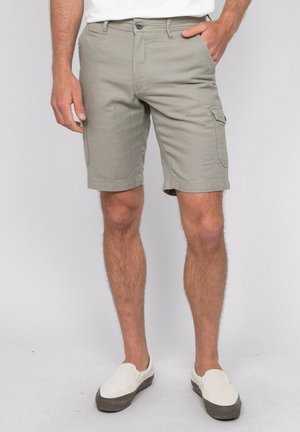 Homme portant un short cargo gris clair, des chaussures blanches sans lacets et une chemise blanche, debout avec une main dans une poche devant un fond uni.