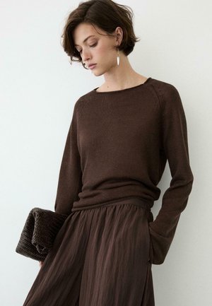 Donna con capelli castani corti che indossa un maglione marrone a maniche lunghe e una gonna fluente, tenendo in mano una pochette marrone testurizzata su uno sfondo bianco semplice.