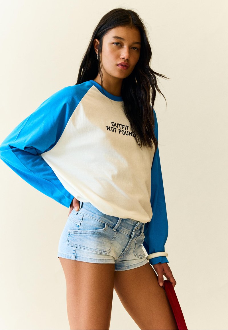 Unge kvinde med langt mørkt hår iført en blå og hvid langærmet skjorte med teksten "OUTFIT NOT FOUND" og lyse denim-shorts, holder en rød genstand.