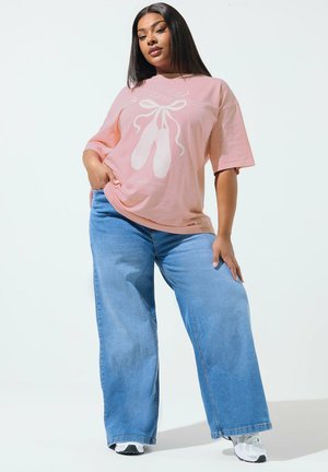 Kobieta o długich, prostych włosach, ubrana w różowy, luźny t-shirt z nadrukiem baletek, szerokie niebieskie dżinsy i białe trampki.