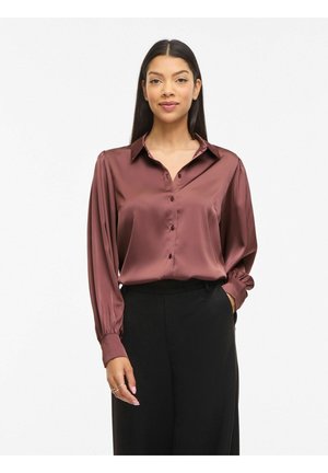 Femeie cu păr lung și întunecat, purtând o bluză strălucitoare mauve cu nasturi și pantaloni negri, stând cu o mână în buzunar pe un fundal alb.