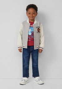 Grijze en witte bomberjack met geborduurde details, rode graphic t-shirt, blauwe jeans en witte schoenen met grijze accenten.