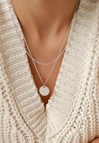 Collane in argento con un pendente rotondo poggiano su un maglione bianco a maglia testurizzata. Il pendente è liscio e circolare, mettendo in risalto il design a strati.