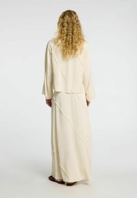 Largo vestido color crema con una chaqueta corta, que presenta un acabado texturizado y detalles de costura diagonal. La tela parece suave y ligera.