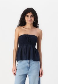 Marinefarget stroppeløs topp med smocked liv og knyte-detalj foran. Utvidet kant laget av lett stoff, komplettert med avslappede denimbukser.