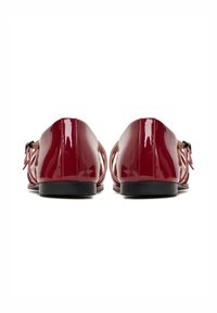 Pair of shiny red patent leather flat shoes with ankle straps and black soles, viewed from the back on a white background.