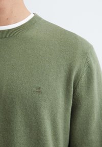 Olijfgroen gebreid truitje met een ronde hals en subtiele textuur, met een kleine geborduurde logo in de onderste linkerhoek.