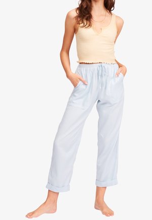 Femme portant un haut beige sans manches en côte et un pantalon bleu clair à cordon avec revers roulés, debout pieds nus, les mains dans les poches.