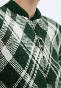 Pull vert et blanc à carreaux avec un col côtelé et une fermeture éclair à l'avant. Il présente une texture tissée et un motif à carreaux variable.