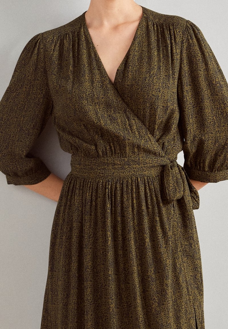 Femme portant une robe portefeuille vert olive avec un petit imprimé léopard noir, des manches trois-quarts bouffantes et une ceinture nouée à la taille.