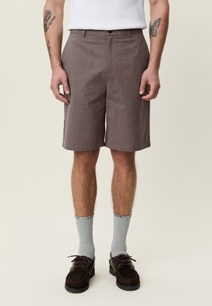 Homme portant un short marron jusqu'aux genoux, des chaussettes grises, des chaussures marron foncé et un débardeur blanc, debout devant un fond clair.