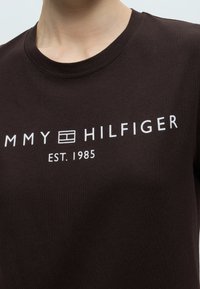 Κοντινό πλάνο ενός σκούρου καφέ T-shirt με λευκό κείμενο "Tommy Hilfiger" και λογότυπο και "EST. 1985" τυπωμένα στο στήθος.