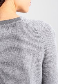 Gros plan d'une personne portant un pull en tricot côtelé gris clair, montrant l'arrière et le côté du cou ainsi que la zone de l'épaule.