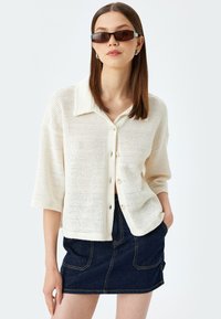 Cremefarvet, løst strikket cardigan med krave og knaplukning, parret med en mørk denim mini-skirt. Model iført solbriller.
