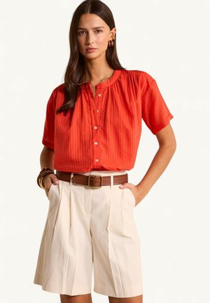 Donna con camicetta rossa a maniche corte con bottoni, pantaloncini beige plissettati a vita alta e cintura marrone, in piedi con le mani nelle tasche.