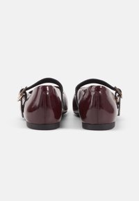 Burgundy kiiltonahasta valmistetut kengät, joissa on pyöreä kärki, takanauhallinen muotoilu ja musta säädettävä hihna. Litteä pohja, jossa on minimaalinen korko.