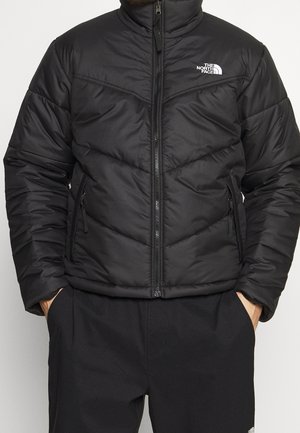 Homme portant une veste matelassée noire North Face, zippée, mains dans les poches, sur un fond clair uni.