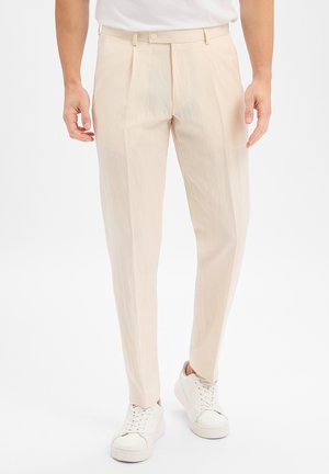 Man wearing cream-colored pleated trousers and white sneakers standing against a plain background.