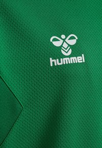 Groene sporttrui gemaakt van textiel met een ademend vermogen, met daarop een wit logo met een gestileerde bij en het woord "hummel" eronder.