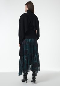 Zwarte trui met een gedrapeerde achterkant over een groen en zwart patroon in lagen rok, gecombineerd met zwarte hoge hakken. Glad en textuurstoffen.