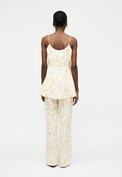 Γυναίκα με σκούρο δέρμα και μαλλιά σε κότσο, που φοράει άσπρο παντελόνι με φαρδιά μπατζάκια και ένα αντίστοιχο αμάνικο τοπ με διακριτικό κίτρινο floral σχέδιο, γυρισμένη προς τα πίσω.