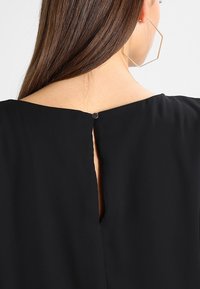 Prenda negra con un diseño de espalda con ojal asegurado con un botón redondo. Textura suave con una silueta minimalista.