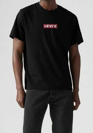 Black short-sleeve T-shirt with red and white "Levi's" logo worn by person with dark jeans against plain background.