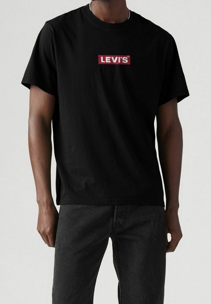 T-shirt noir à manches courtes avec logo "Levi's" rouge et blanc, porté par une personne en jean foncé sur un fond uni.