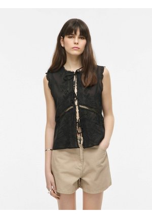 Tânără cu păr lung și castaniu poartă un top negru fără mâneci cu șireturi și pantaloni scurți bej, stând cu o mână în buzunar.