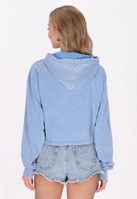 Felpa corta azzurro chiaro con maniche lunghe e cappuccio con cordino, abbinata a pantaloni shorts in denim con orlo sfrangiato. Texture del tessuto morbida.