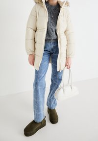 Chaqueta de plumas beige con capucha de piel sintética, suéter gris texturizado, jeans azulados con detalles, y botas de plataforma verde oscuro; bolso blanco.
