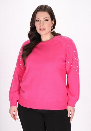 Helles pinkes Sweatshirt mit langen Ärmeln, Perlenverzierungen an den Schultern, gerippten Bündchen und Saum sowie einer lockeren Passform. Kombiniert mit schwarzen Hosen.