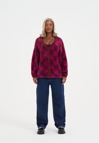 Měkký kostkovaný cardigan v odstínech magenty a burgundské červené. Výstřih do V s knoflíkovým zapínáním. Kombinováno s tmavými džíny s širokými nohavicemi a robustními černými botami.