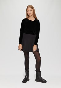 Schwarzes Samt-Oberteil mit langen Ärmeln und quadratischem Ausschnitt, kombiniert mit einem schwarzen, plissierten Rock und schwarzen Ankle Boots mit robuster Sohle.