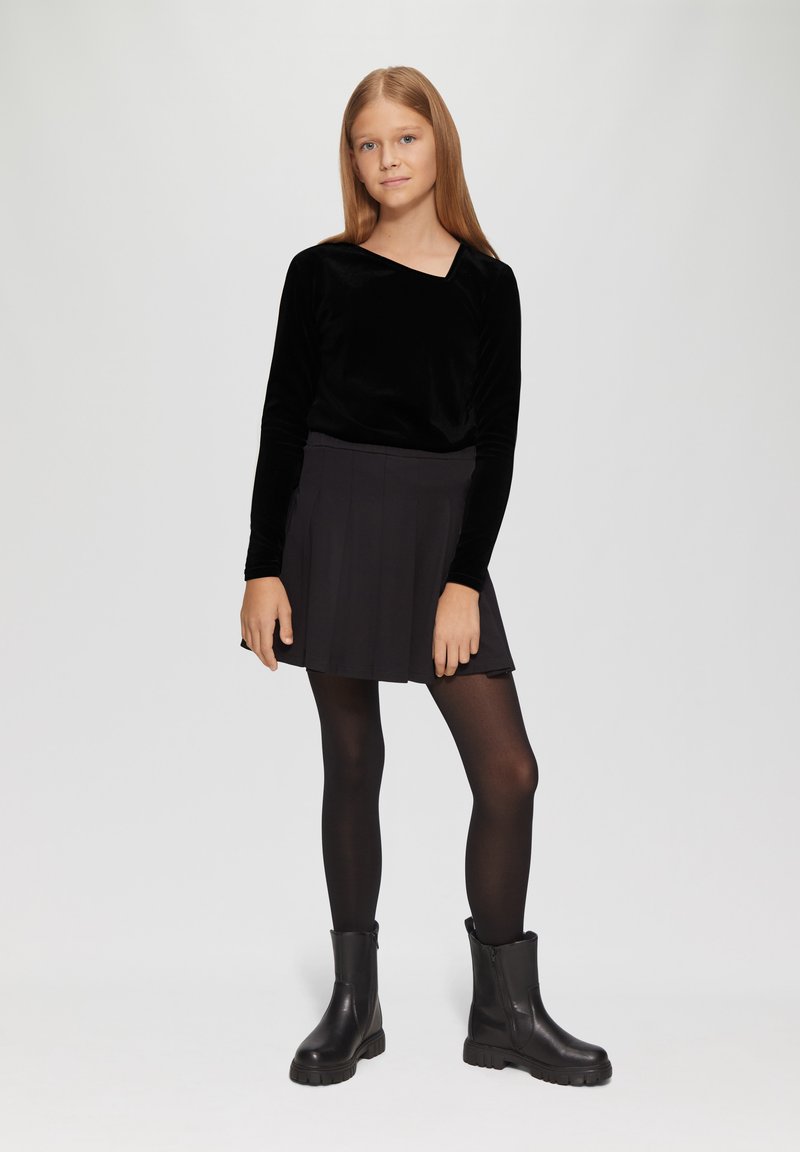 Schwarzes Samt-Oberteil mit langen Ärmeln und quadratischem Ausschnitt, kombiniert mit einem schwarzen, plissierten Rock und schwarzen Ankle Boots mit robuster Sohle.