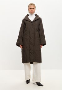 Brun quiltet lang jakke med hætte, sidelommer og brede ærmer, båret over en hvid turtleneck og lyse bukser. Sorte spidse hæle.