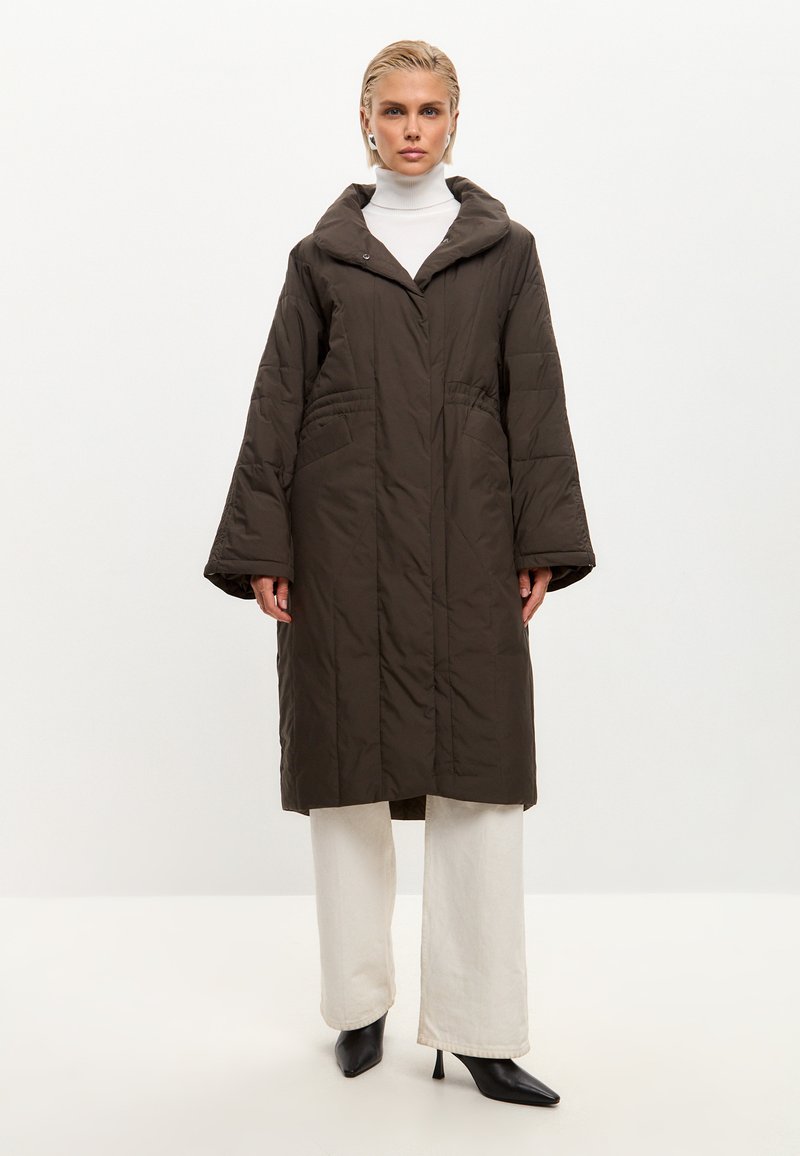 Brun quiltet lang jakke med hætte, sidelommer og brede ærmer, båret over en hvid turtleneck og lyse bukser. Sorte spidse hæle.