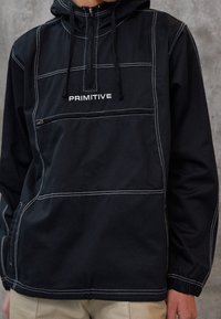 Veste à capuche noire avec coutures blanches et texte "PRIMITIVE" sur la poitrine, poche zippée sous le texte, portée par une personne portant un pantalon marron clair.