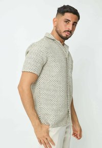 Chemise à manches courtes en crème avec un motif géométrique bleu et noir, dotée d'un col boutonné et d'un tissu léger et texturé.