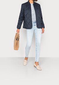 Marinefarvet quiltet jakke med rund krave, hvid mønstret top, lyseblå skinny jeans og beige loafers, parret med en tan håndtaske.