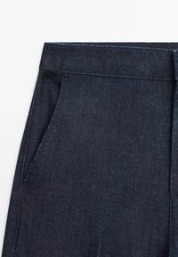 Gros plan d’un pantalon en jean bleu foncé montrant la ceinture et le détail de la poche latérale sur fond blanc.