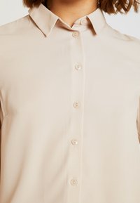 Gros plan d'une personne portant une chemise beige à boutons avec un col classique et cinq boutons visibles sur le devant.