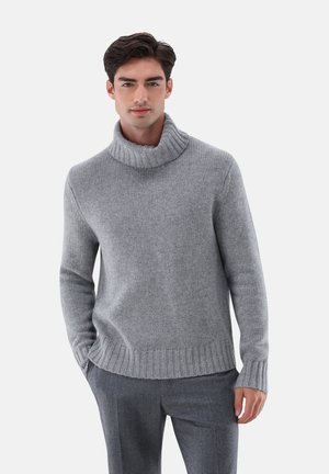 Grauer Strickpullover mit hohem, geripptem Kragen, langen Ärmeln und lockerer Passform. Kombiniert mit grauen Hosen. Weiche Textur und nahtloses Design.