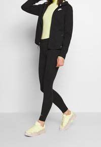 Svart zip-up jacka över en gul polotröja, tillsammans med svarta leggings och ljus gul-grå sneakers med snördesign och texturerade sulor.