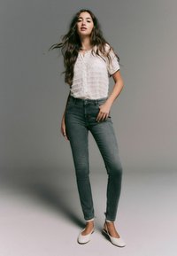 Witte, gestructureerde blouse met korte mouwen, hoogwaardige donkerblauwe jeans en witte flats. Het model staat met één hand op de heup en toont de outfits.